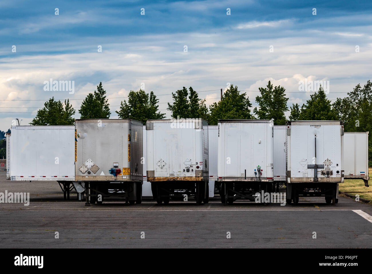 Semi tractor trailers parked in a row with doors closed under a blue cloudy sky with trees Stock Photo