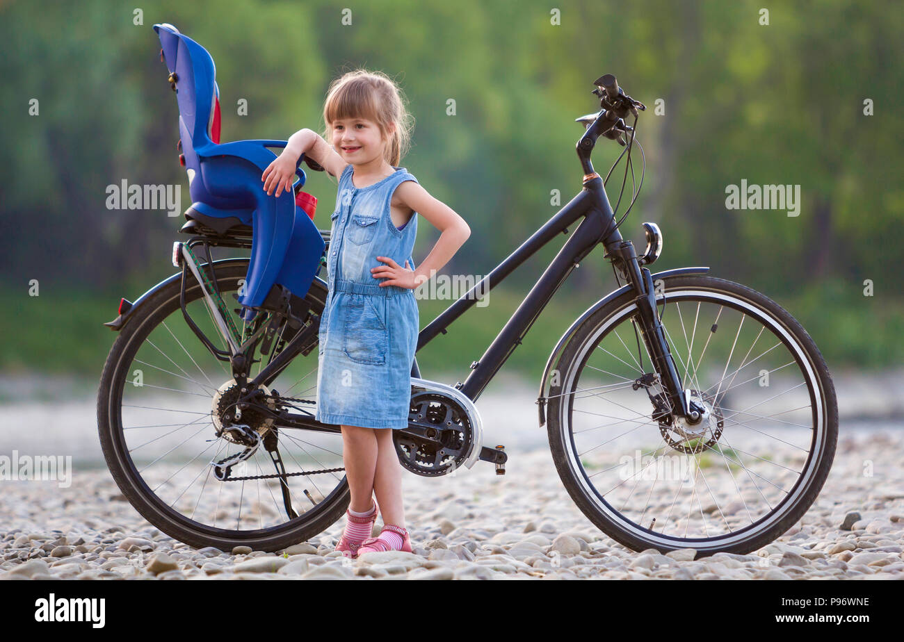 Small pretty blond girl in blue dress standing on pebbles in front of modern bicycle with child seat on blurred green trees background on summer day.  Stock Photo