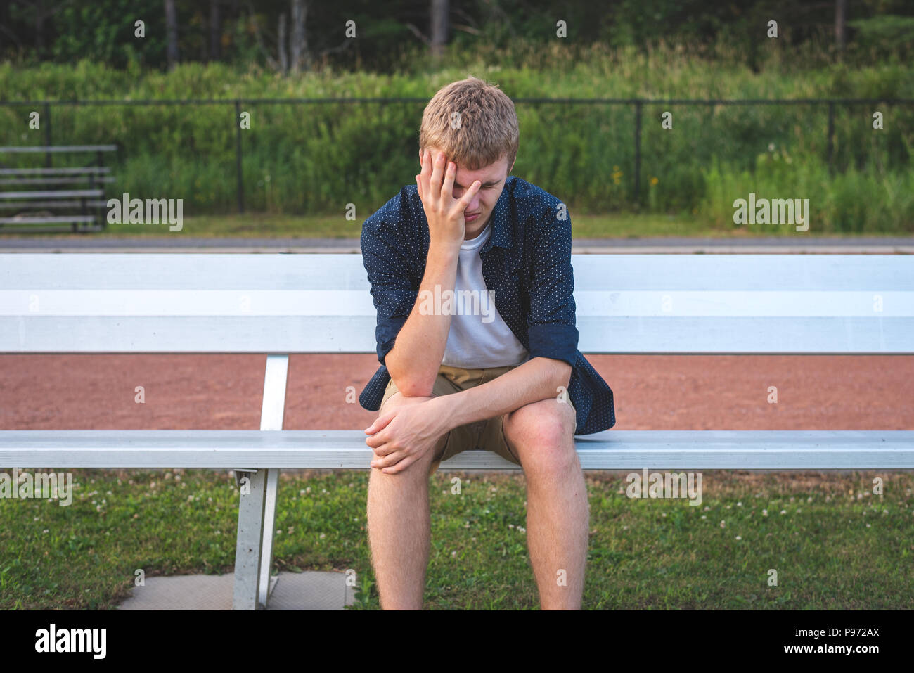 Sad teenager sitting on a school sports field team bench next to a running track. Stock Photo