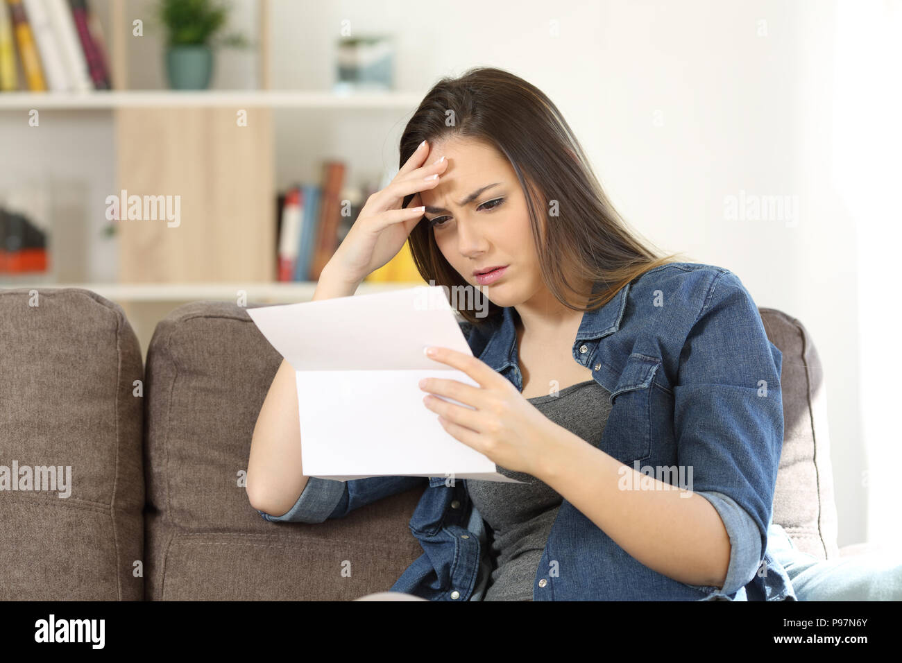 Concerned woman reading bad news in a letter sitting on a couch in the living room at home Stock Photo