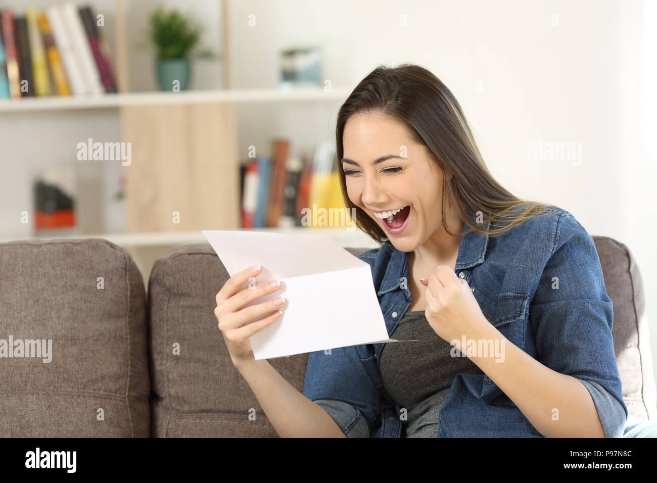 Excited woman reading great news in a letter sitting on a couch in the living room at home Stock Photo