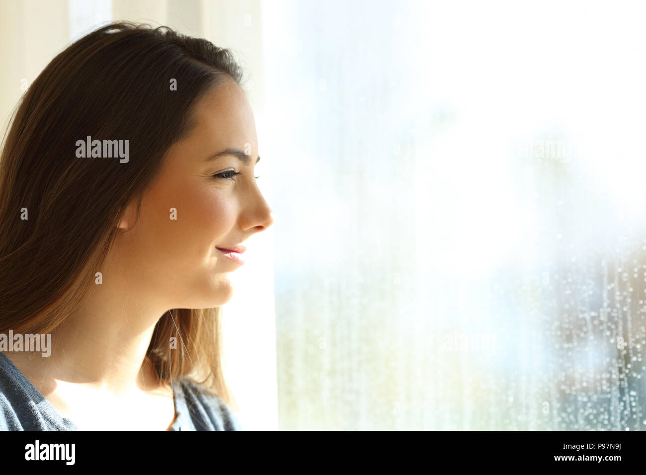 Side view portrait of a happy girl looking at the sun shining through a window after the rain Stock Photo