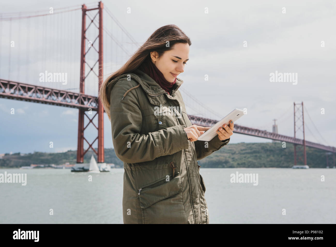 A young beautiful woman uses a tablet to communicate with friends or looks at a map or calls a taxi or something else. Bridge April 25 in Lisbon in Po Stock Photo