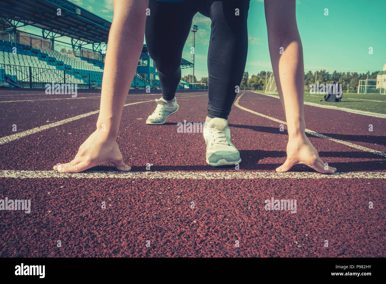Ready to go. Young fitness girl wearing blue top, black tights, sneakers and pony-tail on the starting line of stadium track, preparing for a run. Hea Stock Photo
