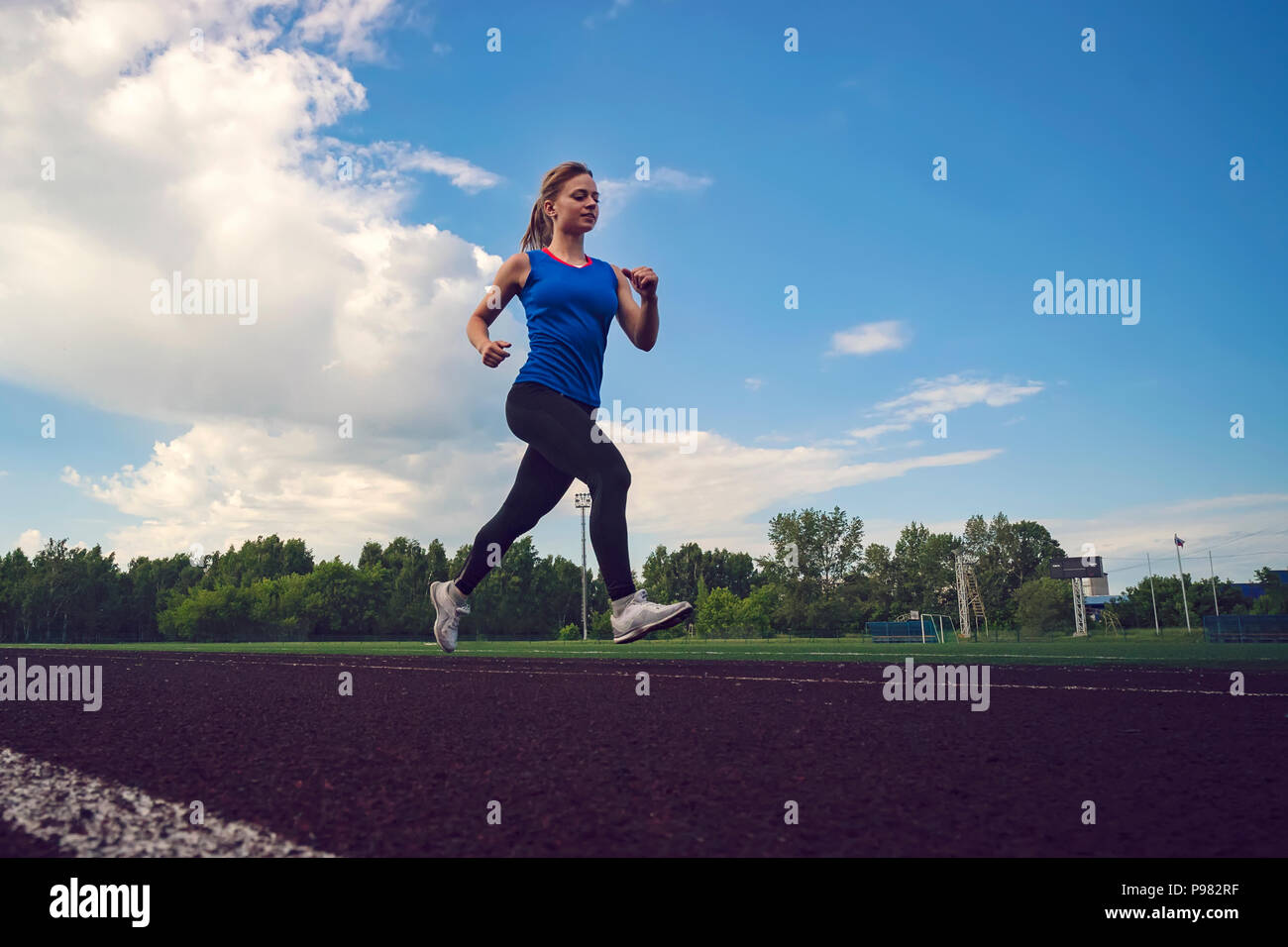 Young woman running during sunny evening on stadium track. A blonde in a blue t-shirt and black leggings runs across the stadium. Stock Photo