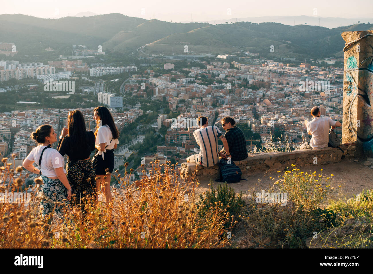 a young group of tourists sit atop Bunquers del Carmel at sunset, a famous lookout over the city of Barcelona, Spain Stock Photo