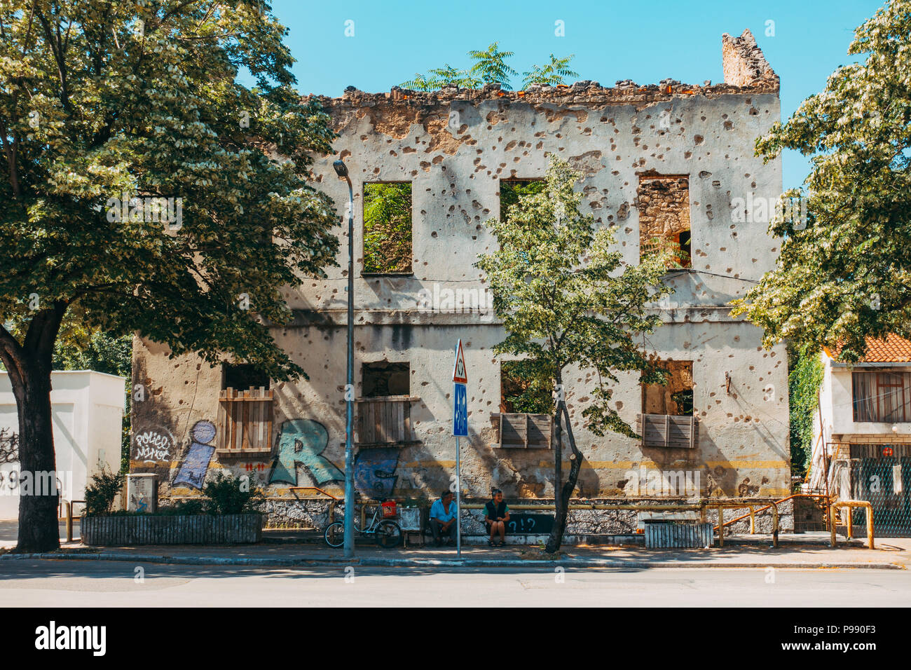 Two locals sit in front of the remains of a artillery shell-ridden building facade, in Mostar, Bosnia and Herzegovina Stock Photo