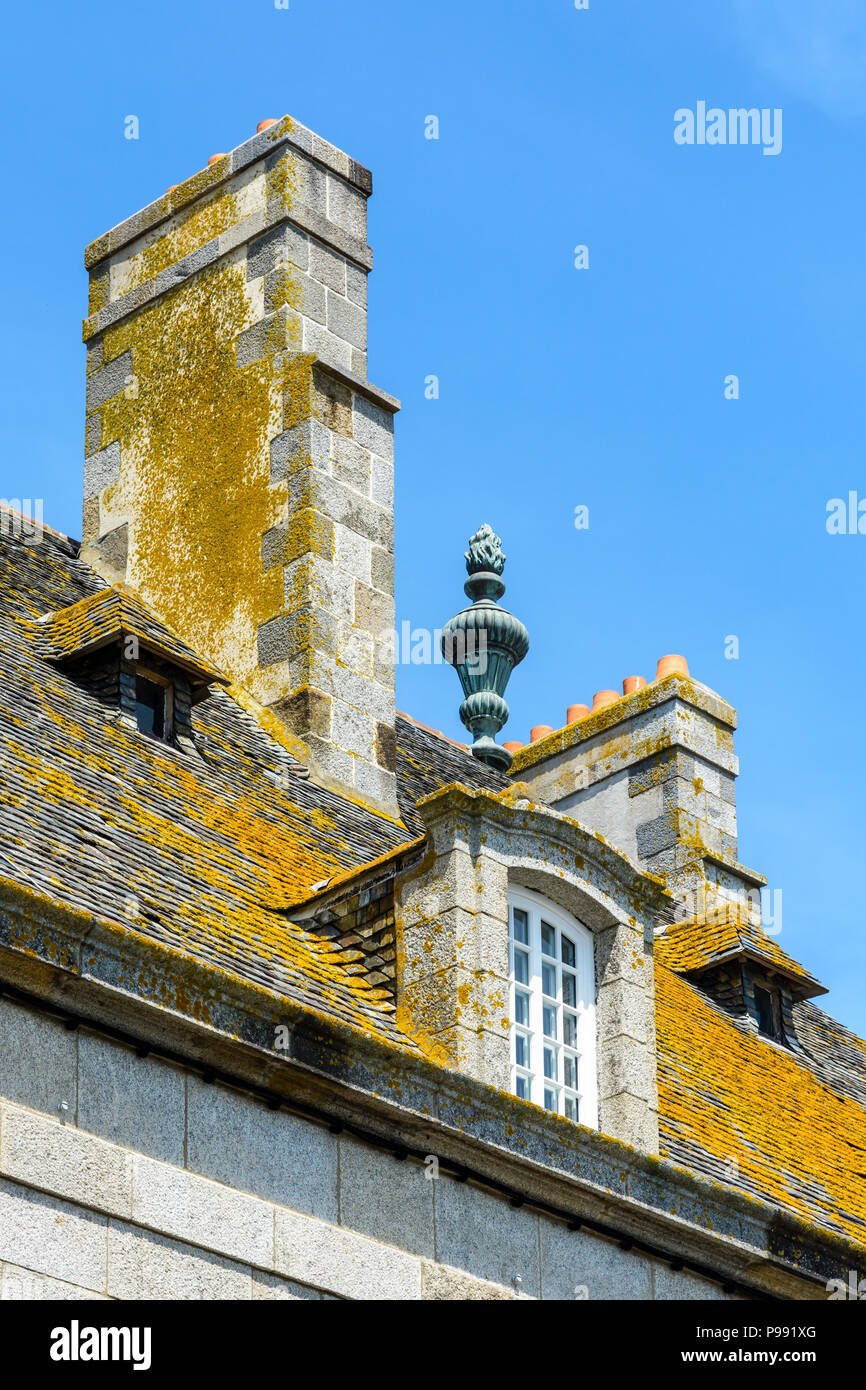 The slate roof covered with lichen, the dormer windows and chimneys of a residential building in the old city of Saint-Malo, France, against blue sky. Stock Photo