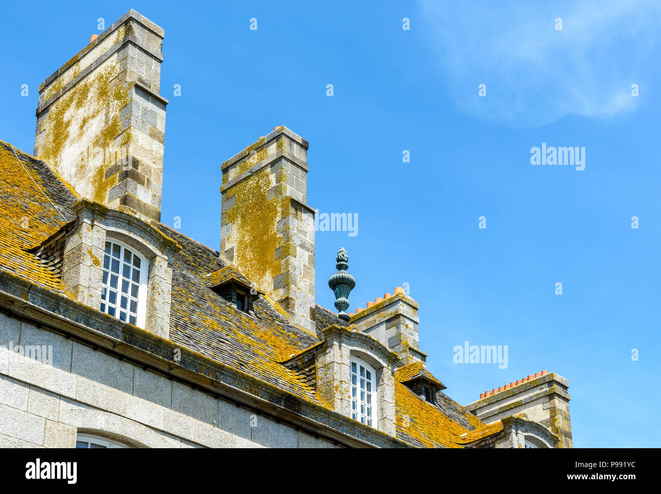 The slate roof covered with lichen, the dormer windows and chimneys of a residential building in the old city of Saint-Malo, France, against blue sky. Stock Photo