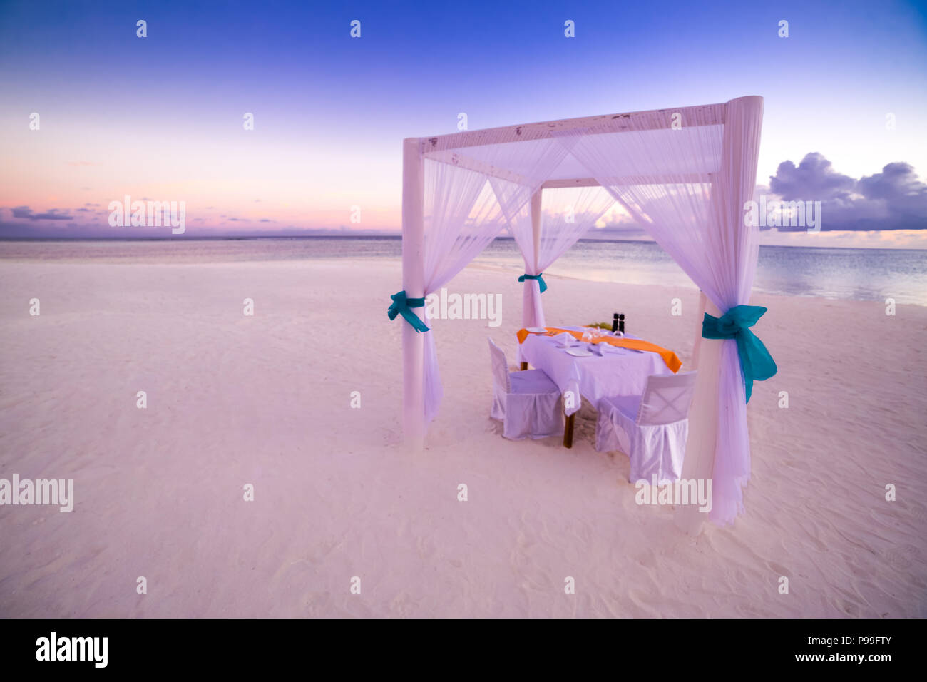 Romantic dinner table set-up for a honeymoon couple on the beach under sunset sky. Exotic wedding and honeymoon concept Stock Photo