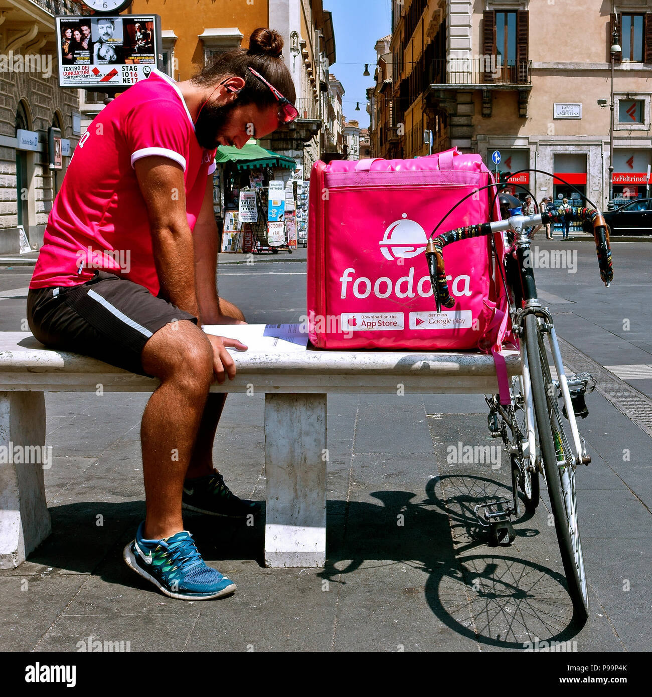 Delivery boy wearing a pink tshirt, resting on a bench and studying while waiting for a delivery request call. Pink delivery courier box. Rome, Italy Stock Photo