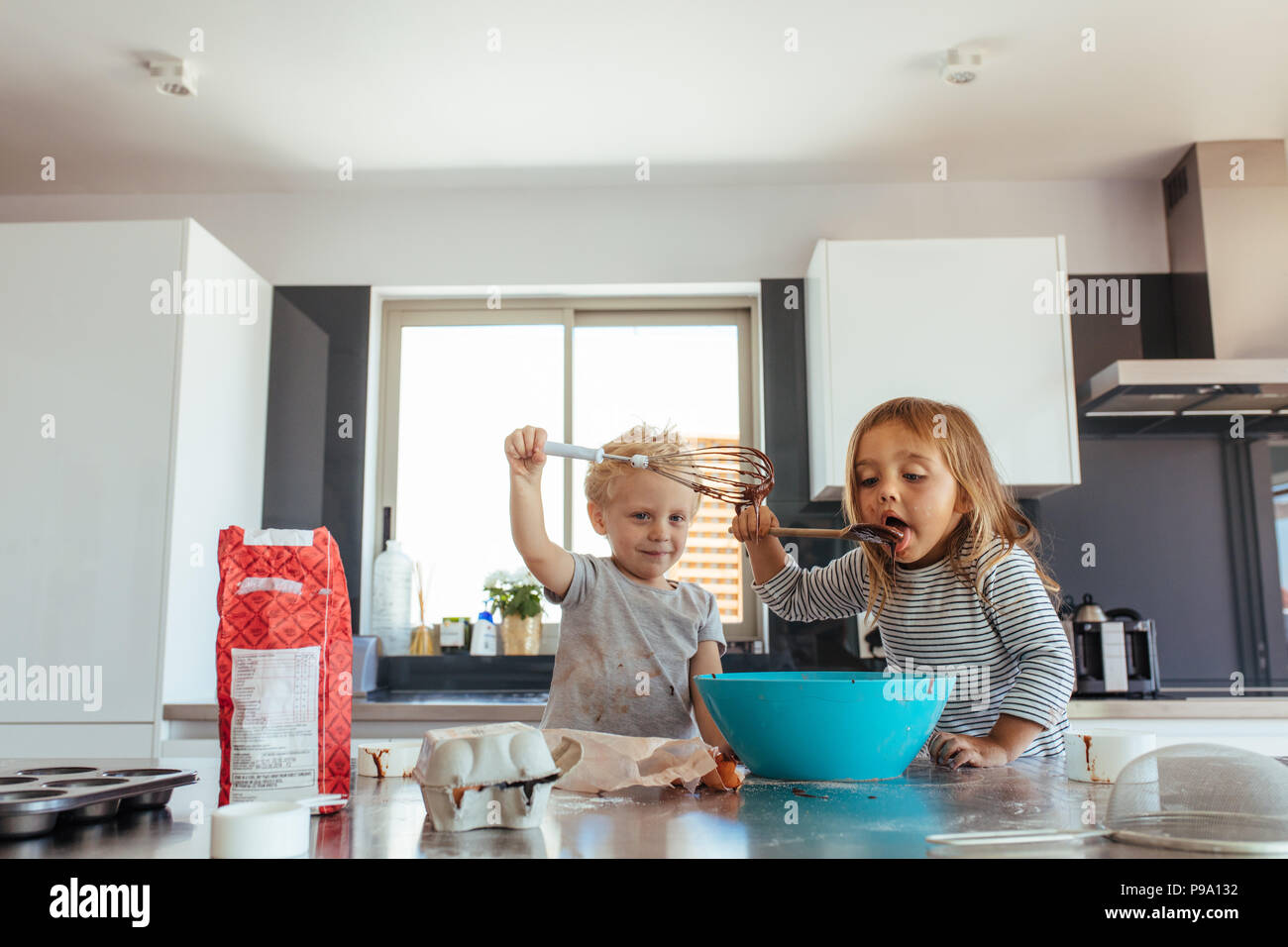 Young girl licking batter from spatula with her brother holding a whisk. Siblings making cake batter in kitchen. Stock Photo