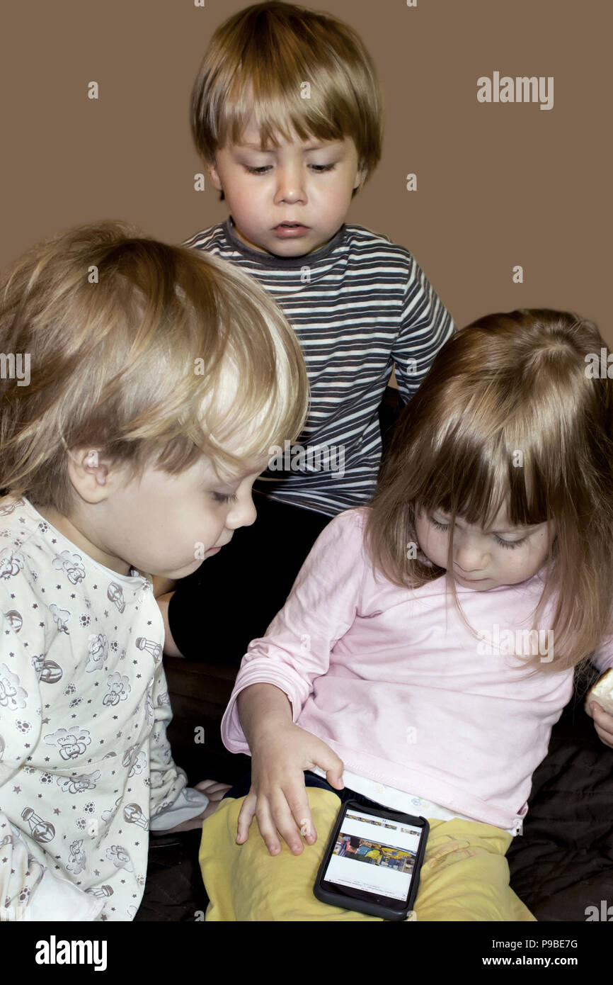 Little cute curious blonde triplets watch cartoons on smartphone indoors, half resaturated image isolated on dark beige Stock Photo