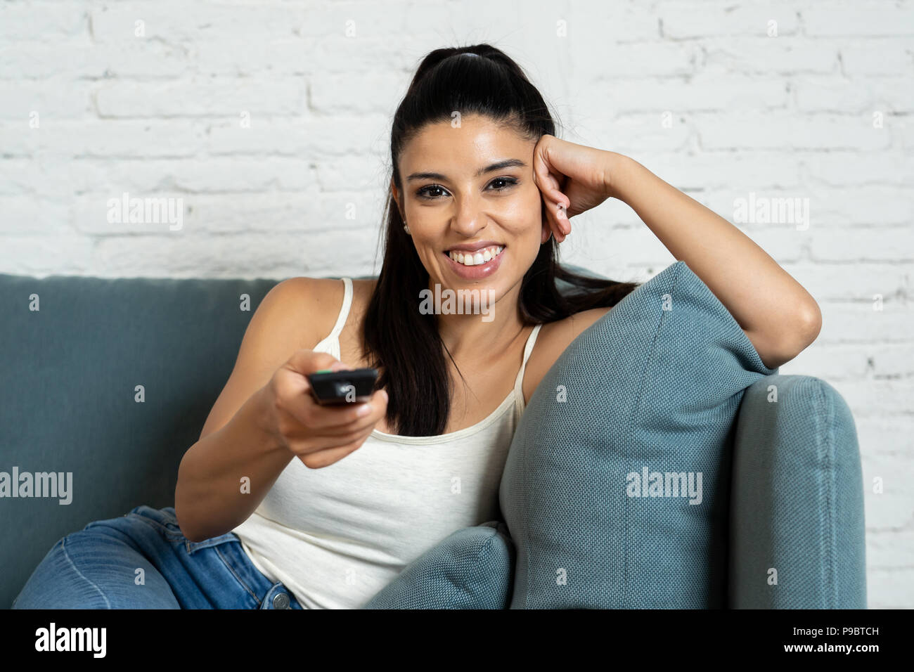 young beautiful Spanish woman in jeans home alone watching television smiling sitting on sofa couch in living room happy and excited enjoying a TV rom Stock Photo