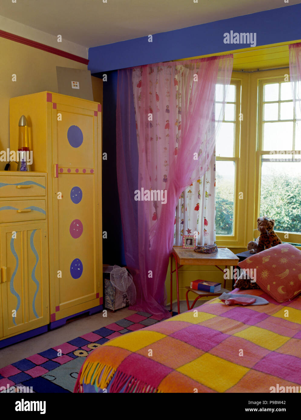Pink net drapes on window in a nineties economy style children's bedroom with yellow painted cupboards Stock Photo