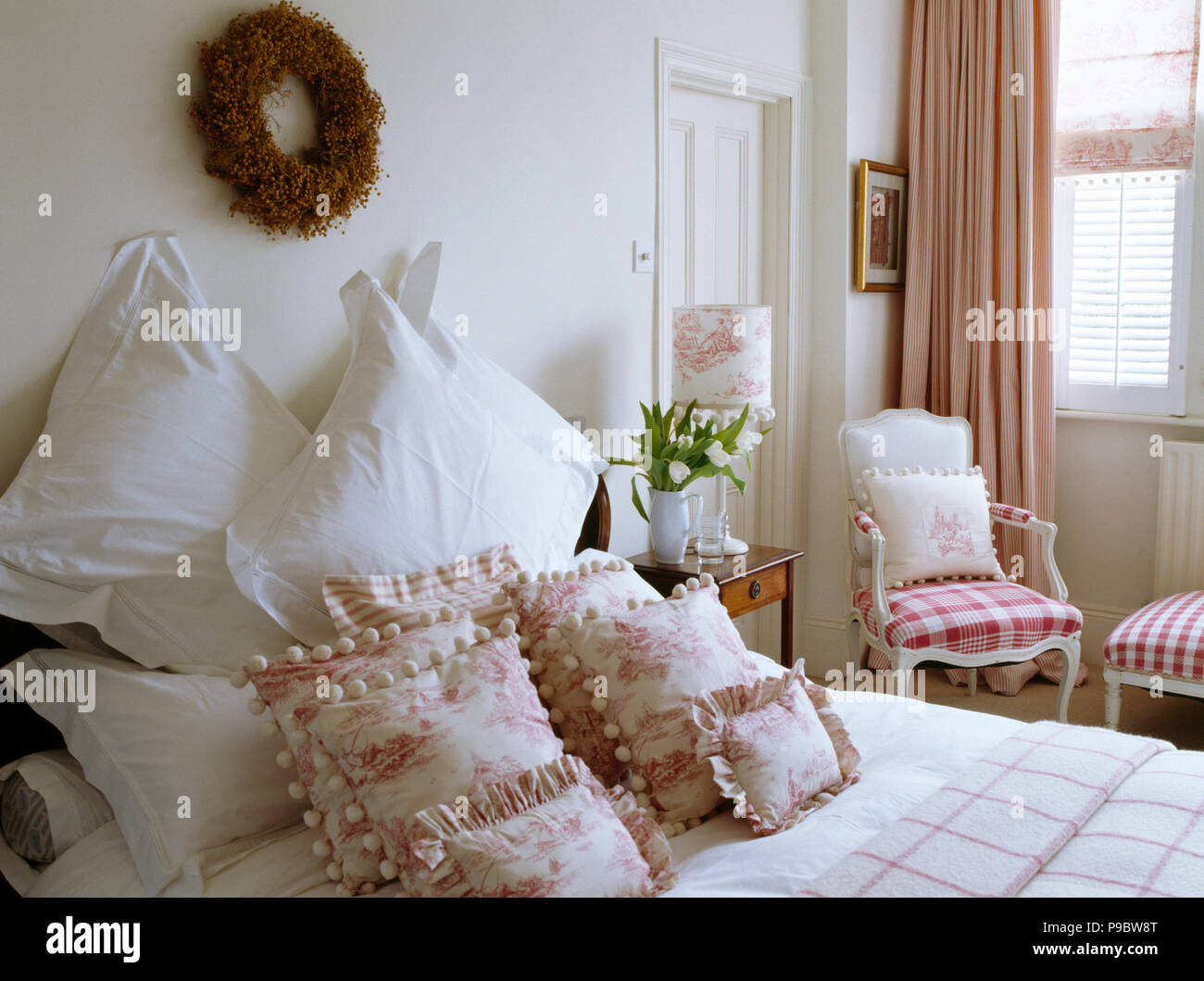 Twig wreath above bed with white pillows and pink+white Toile de Jouy cushions in traditional white bedroom Stock Photo