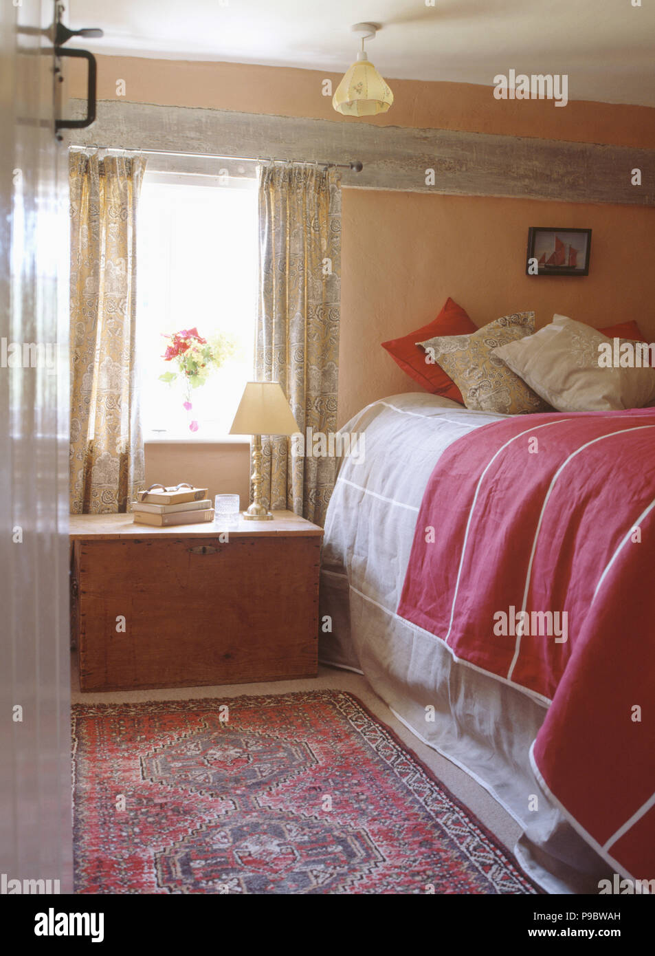 Wooden chest below window in cottage bedroom with cushions and red throw on the bed Stock Photo