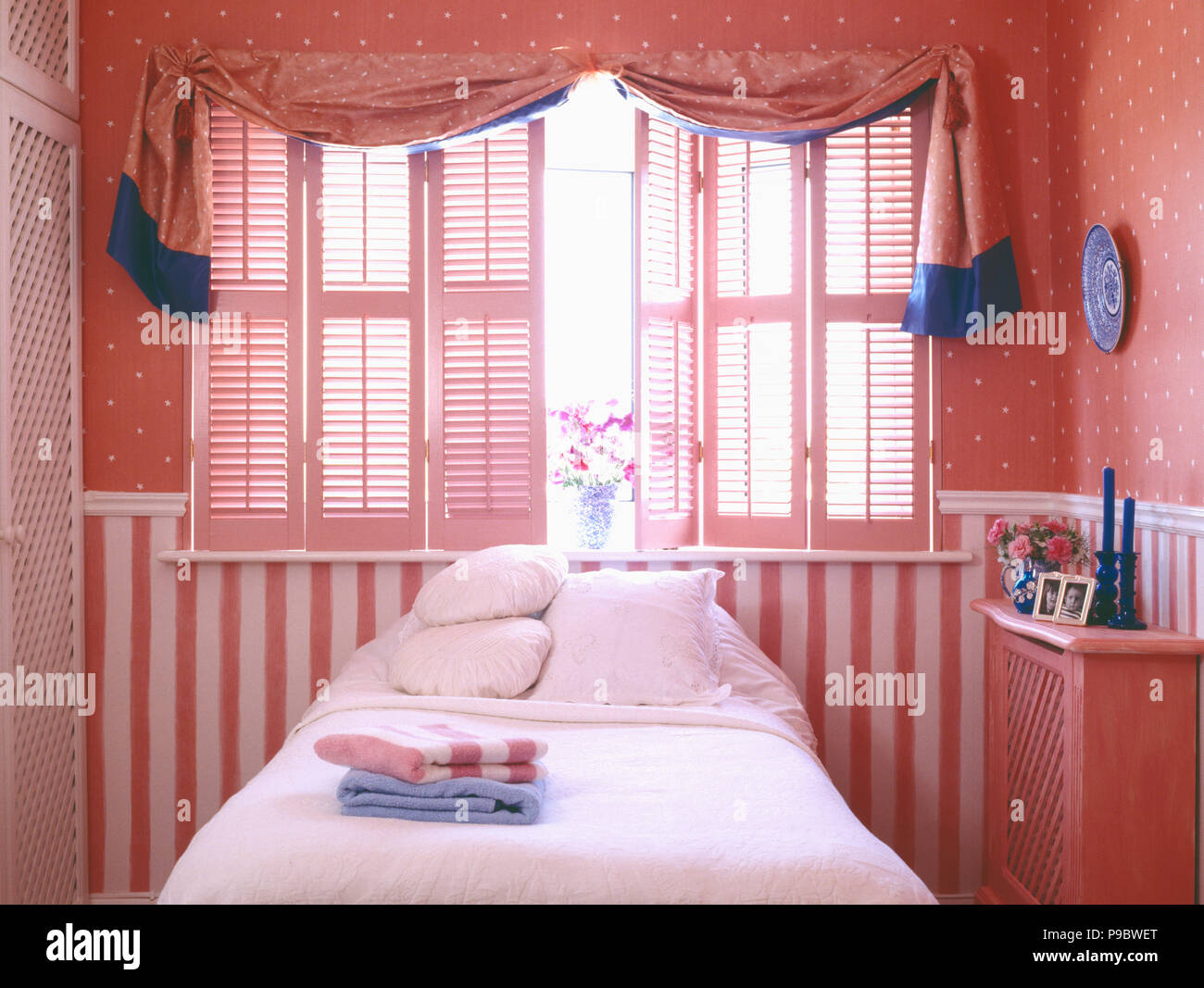 Draped pink fabric over plantation shutters in nineties bedroom with pink striped and spotted wallpaper Stock Photo