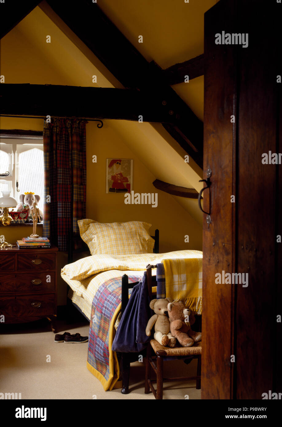 Teddy bears on chair at the foot of a bed with yellow and white checked bed linen in a child's yellow attic bedroom Stock Photo