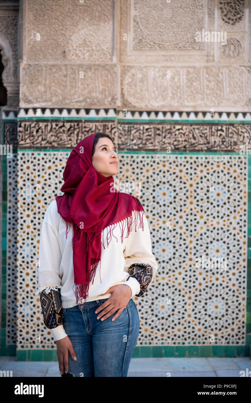 Muslim woman posing and looking up in traditional clothing with red hijab and jeans in front of traditional arabesque decorated wall Stock Photo
