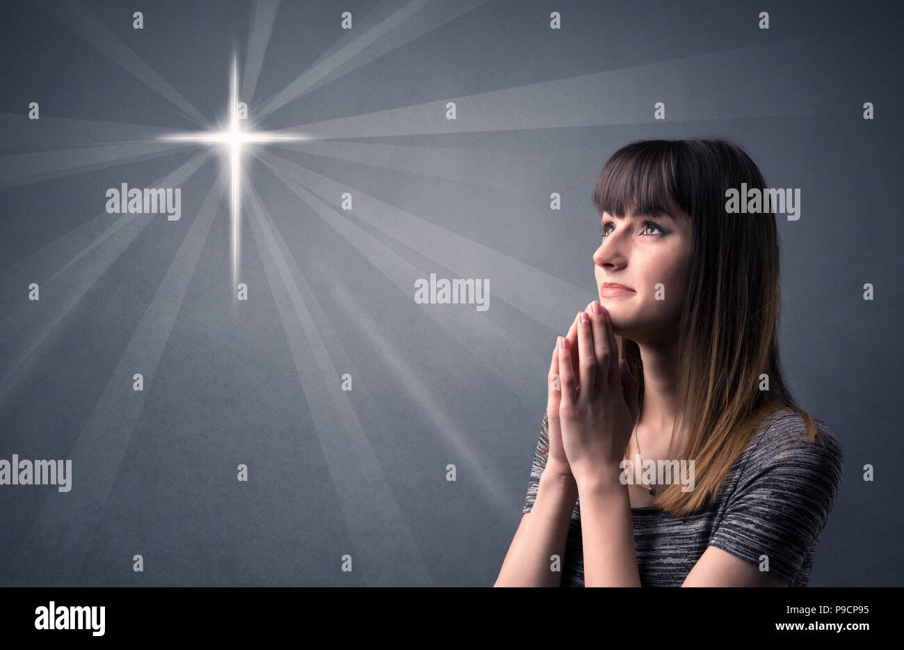 Young woman praying on a grey background with a shiny cross silhouette ...