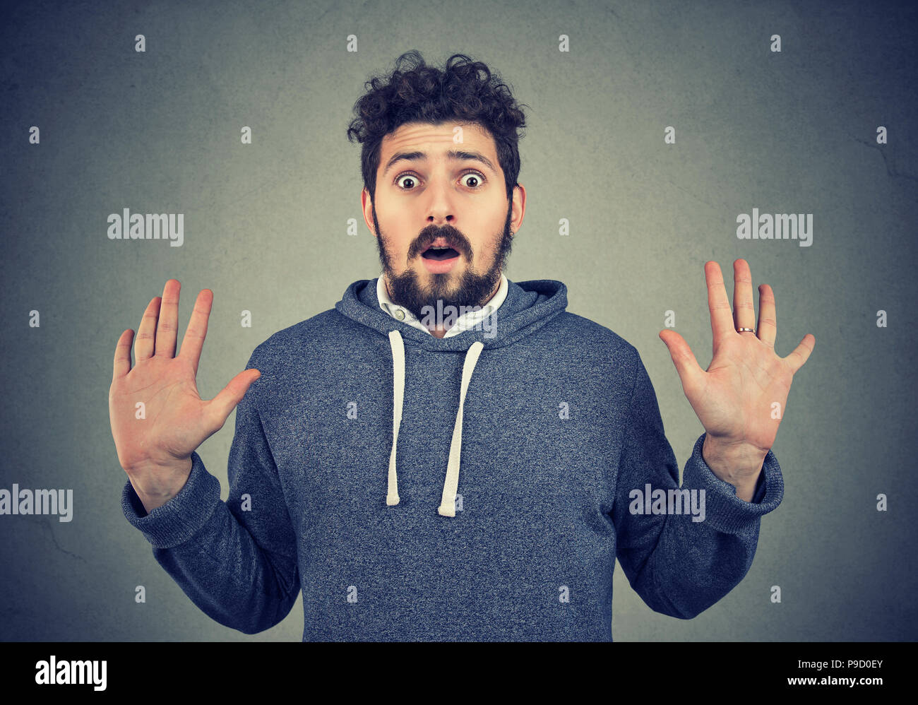 Young bearded man in hoodie looking with fear and astonishment at camera with hands up on gray background Stock Photo