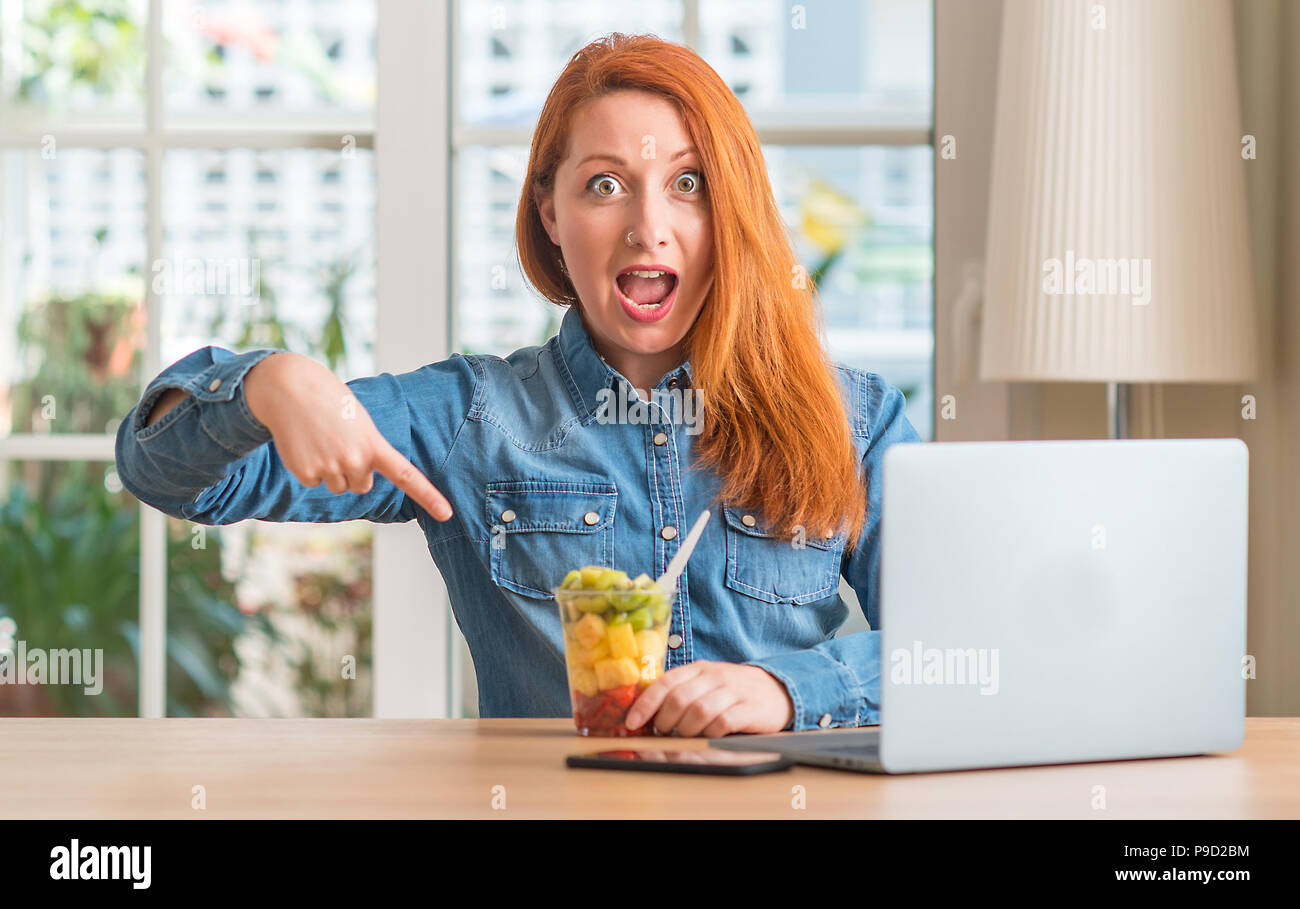 Redhead woman using computer laptop eating fruit at home very happy ...