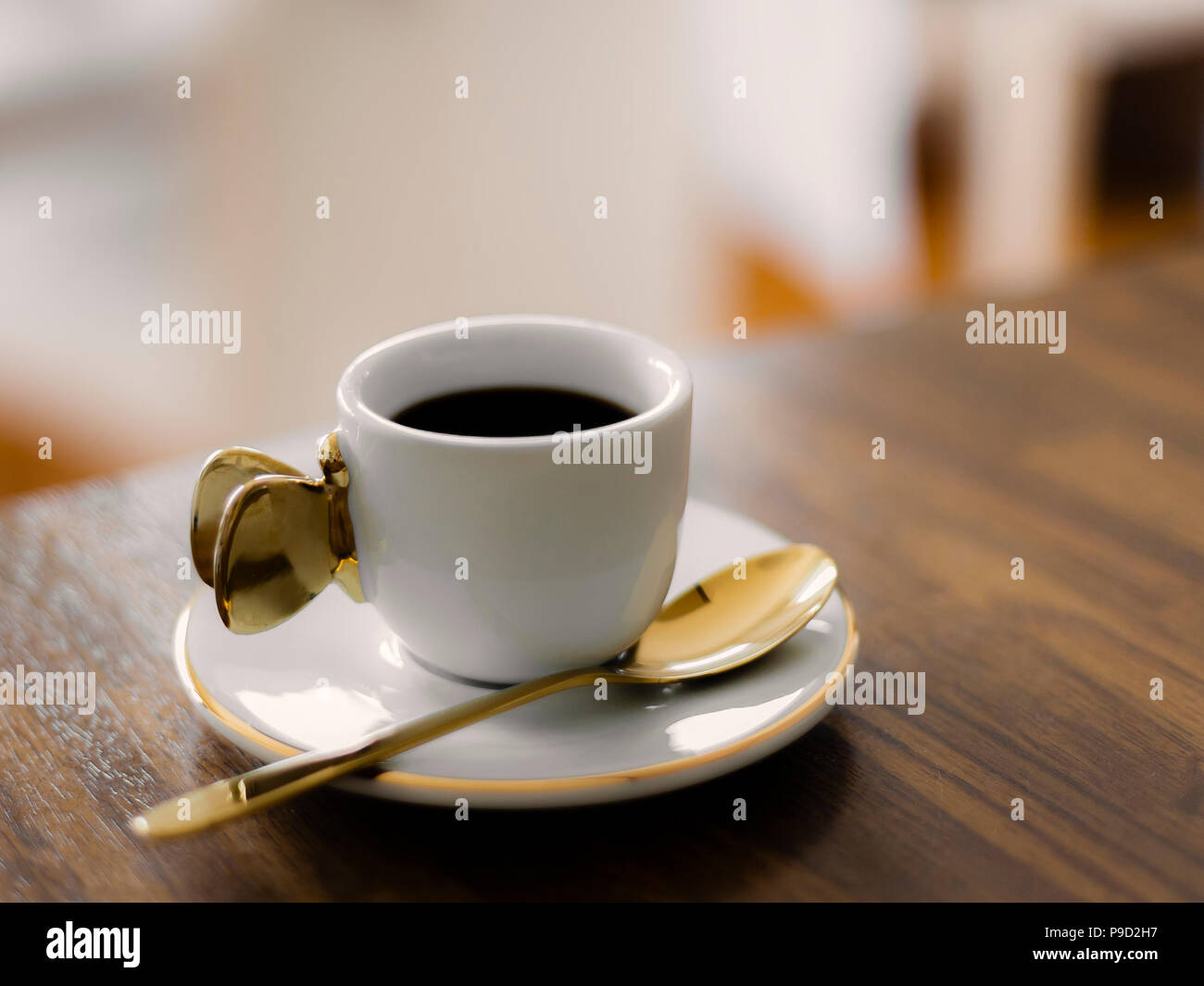 White cup with a golden handle, saucer and spoon, filled with black coffee on a wooden counter. Stock Photo