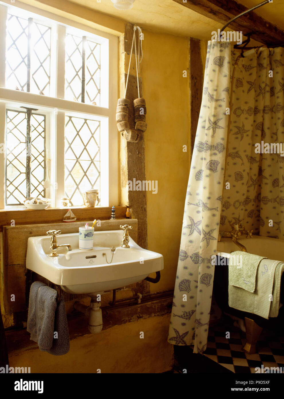 String of cork floats on window above basin in country bathroom with seashore-patterned shower curtain on bath Stock Photo