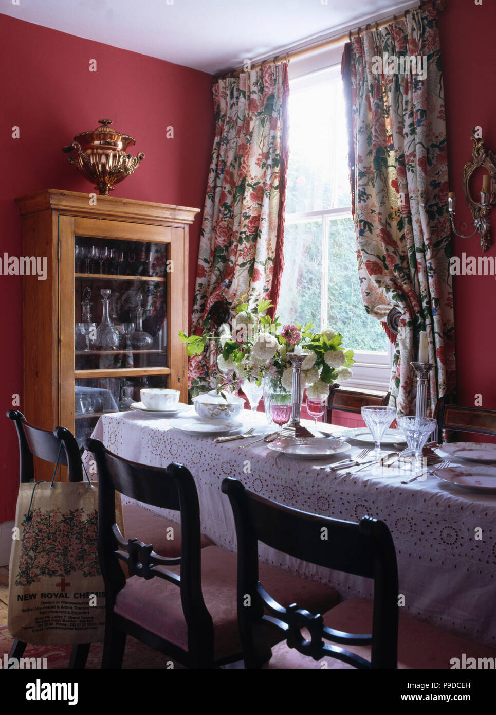 White linen cloth on table set for lunch in Madder-red dining room with floral curtains at the window Stock Photo