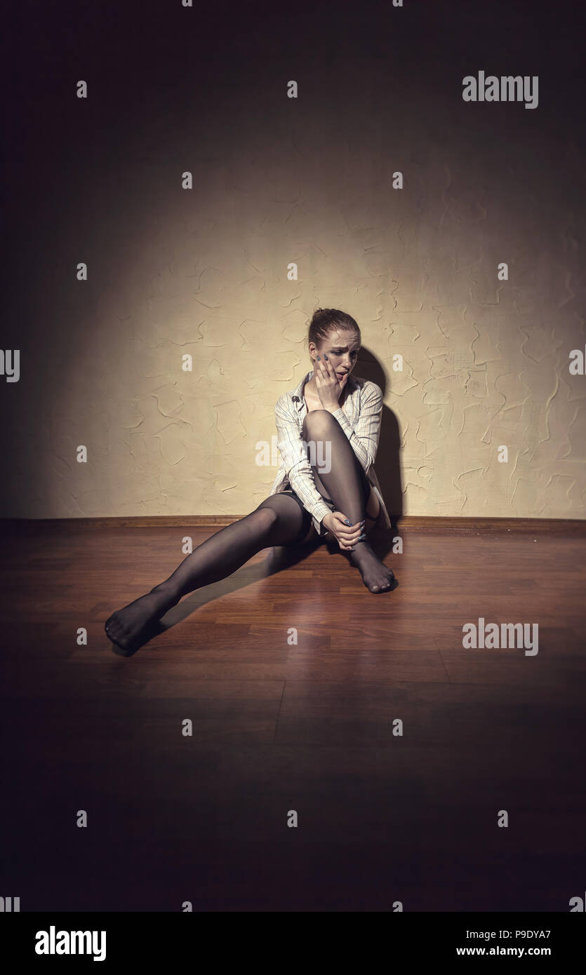 A crying young woman sits alone by the wall in an empty room Stock Photo