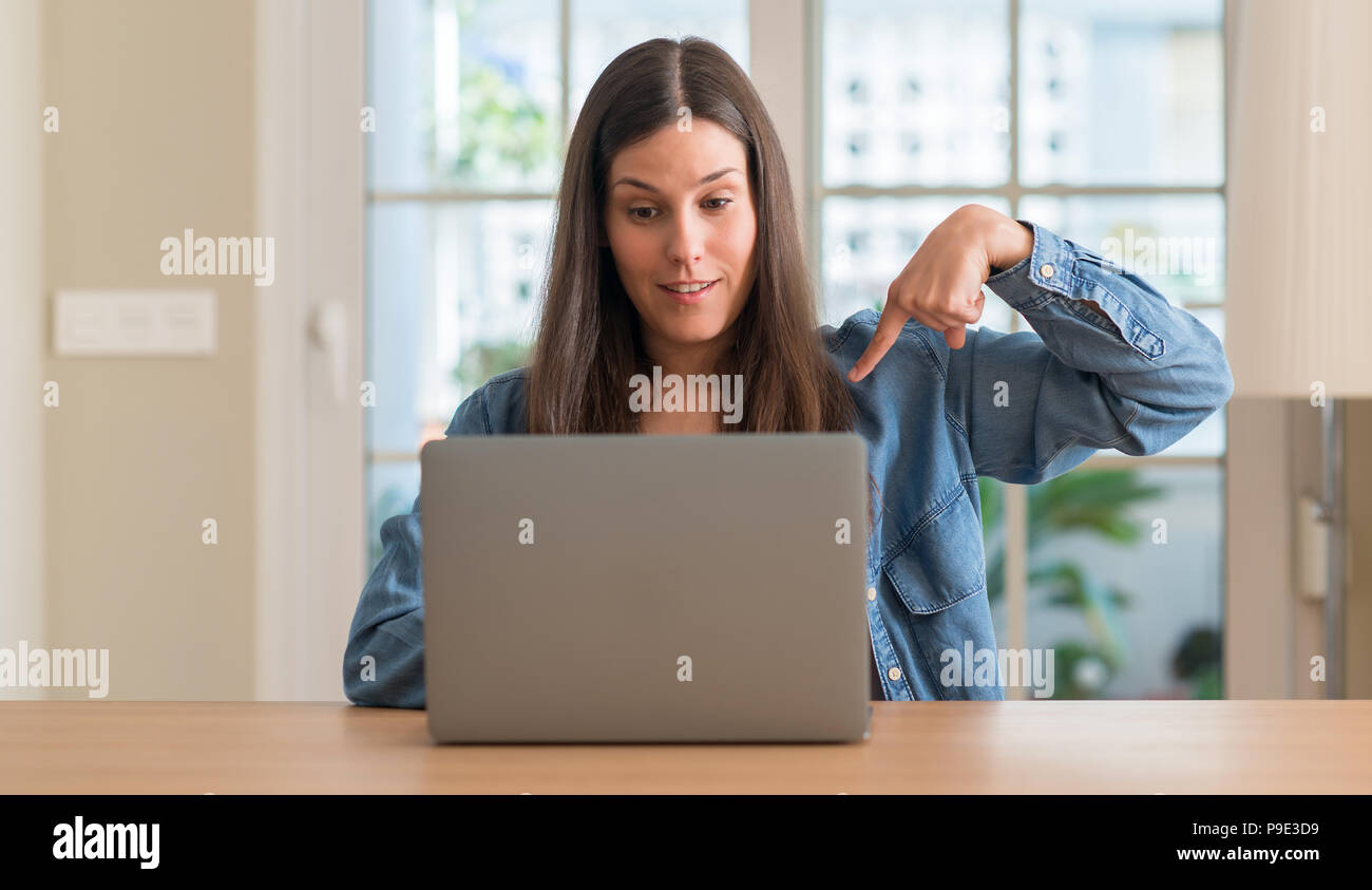 Young woman using laptop at home with surprise face pointing finger to ...