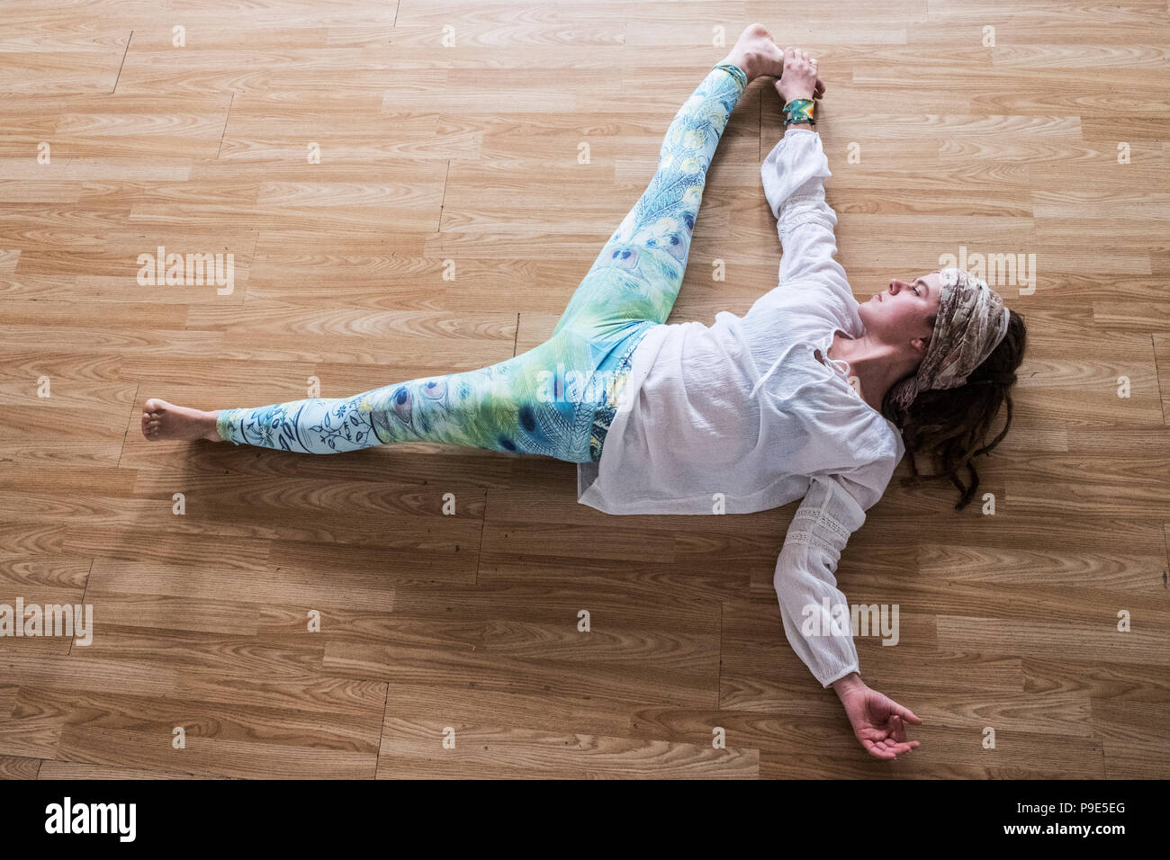 High angle view of young woman wearing headscarf and white blouse lying on her back on hardwood floor, doing yoga. Stock Photo