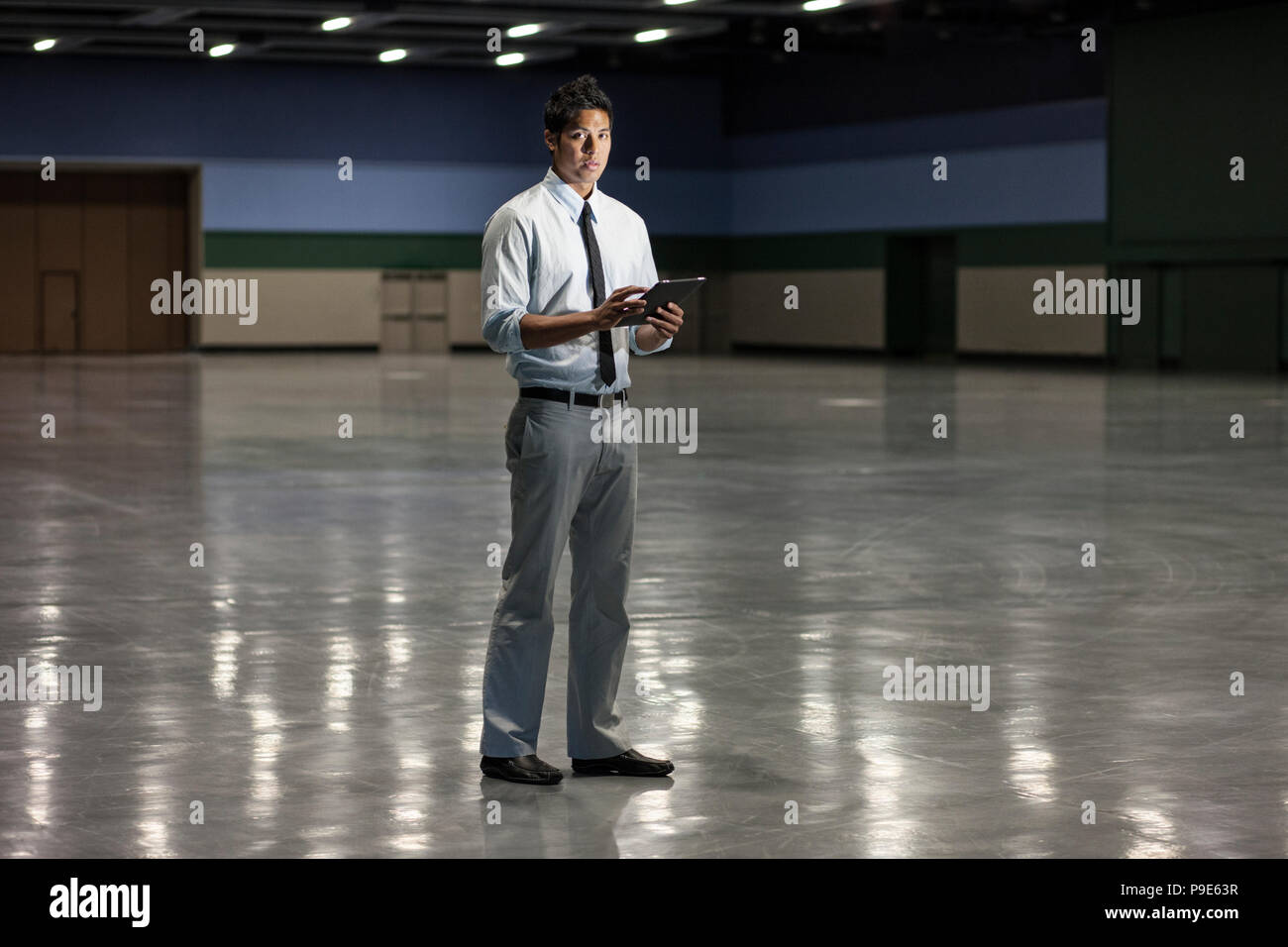 Portrait of a businessman holding a notebook computer and standing in a dimly lit and dark exhibition area in a convention center. Stock Photo