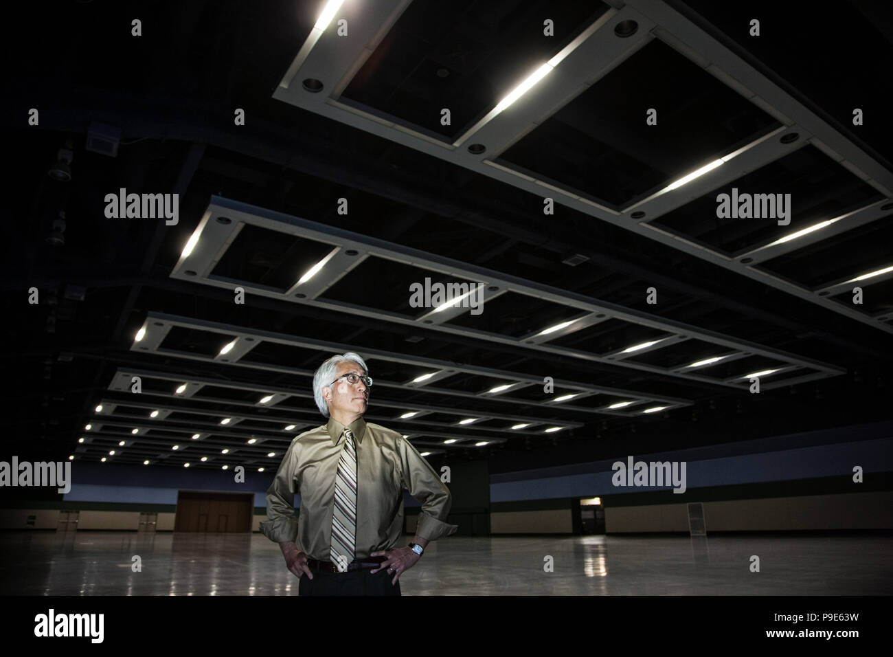An Asian businessman standing in a dimly lit and dark exhibition area in a convention center. Stock Photo