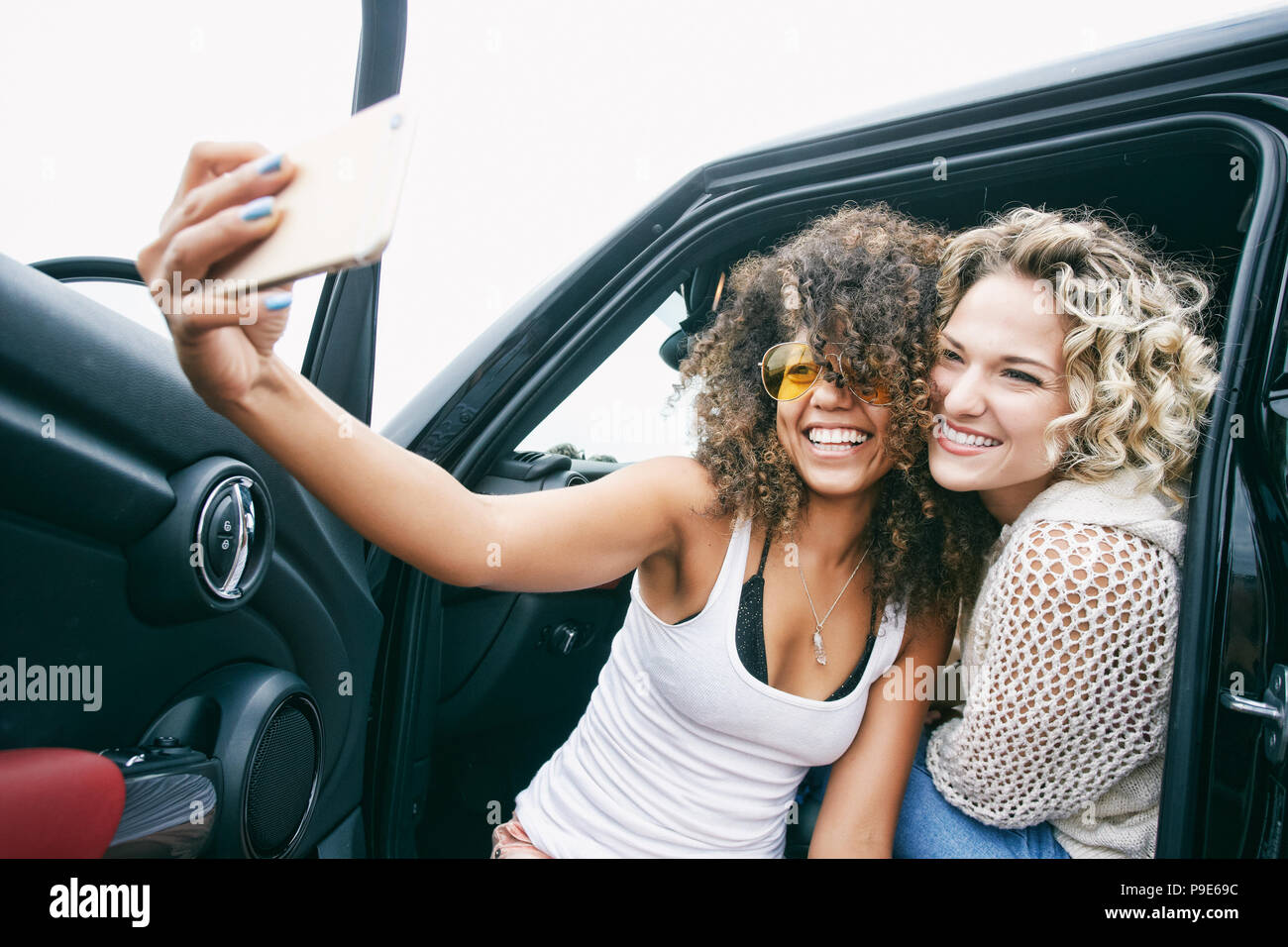 Portrait of two smiling young women with blond and brown curly hair sitting in car, taking selfie with mobile phone. Stock Photo