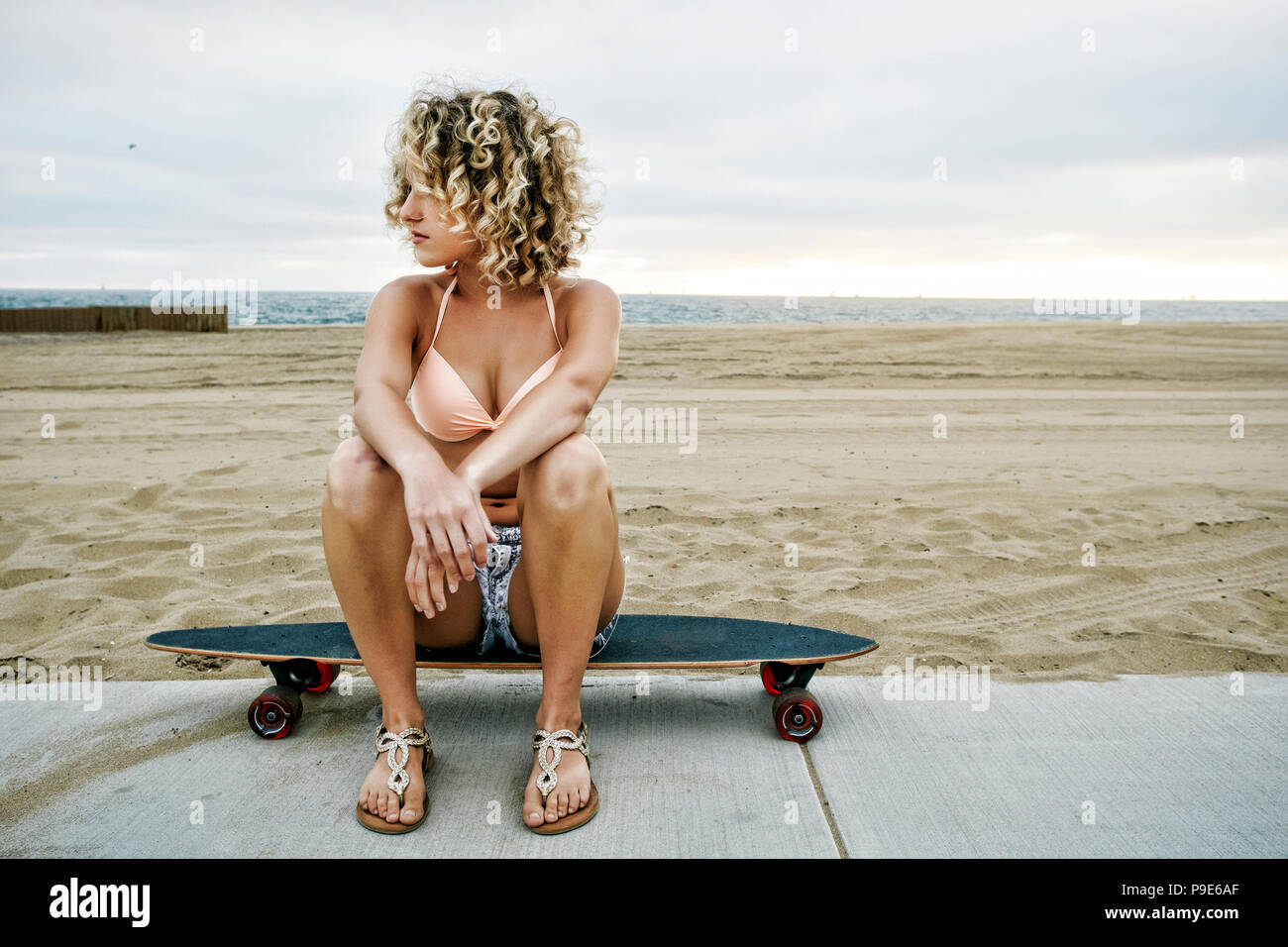 Young woman with curly blond hair wearing pink bikini sitting on skateboard  on sandy beach Stock Photo - Alamy