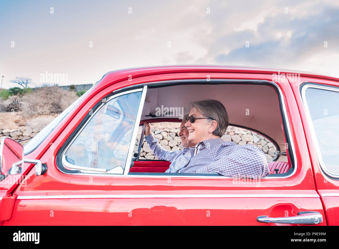 Elderly couple with hat, with glasses, with gray and white hair, with casual shirt, on vintage red car on vacation enjoying time and life. With a chee Stock Photo