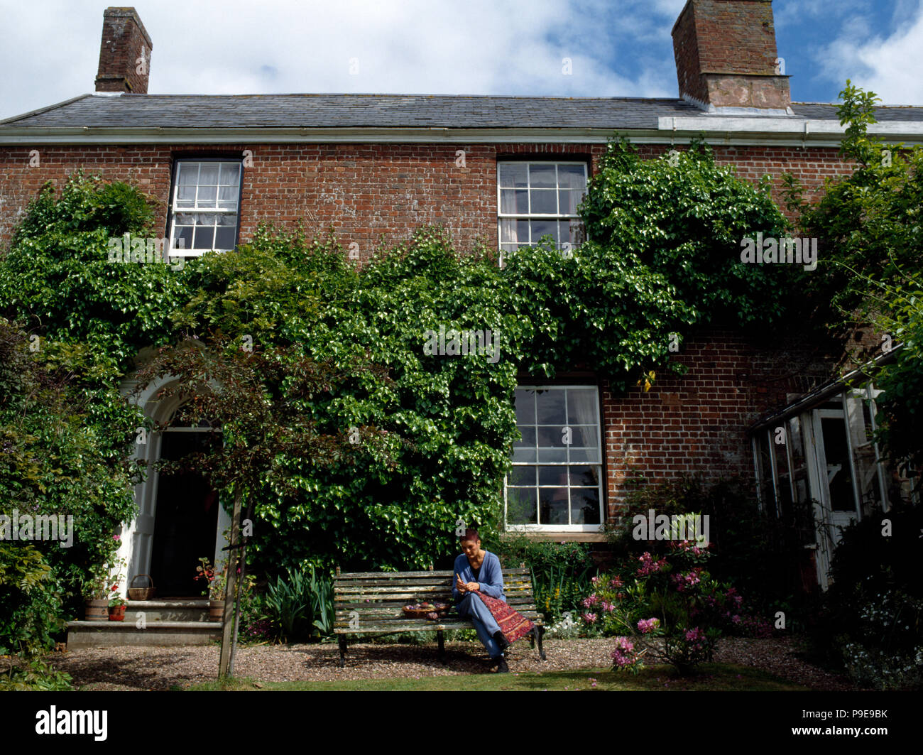 Exterior of a Victorian house with Virginia creeper on the walls and a woman sitting on bench and sewing Stock Photo