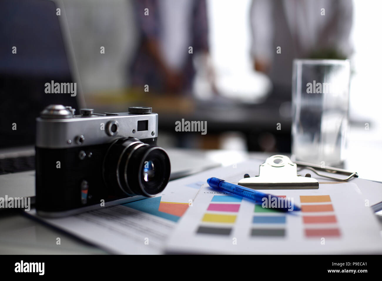 Laptop and camera on the desk in office, people standing on background