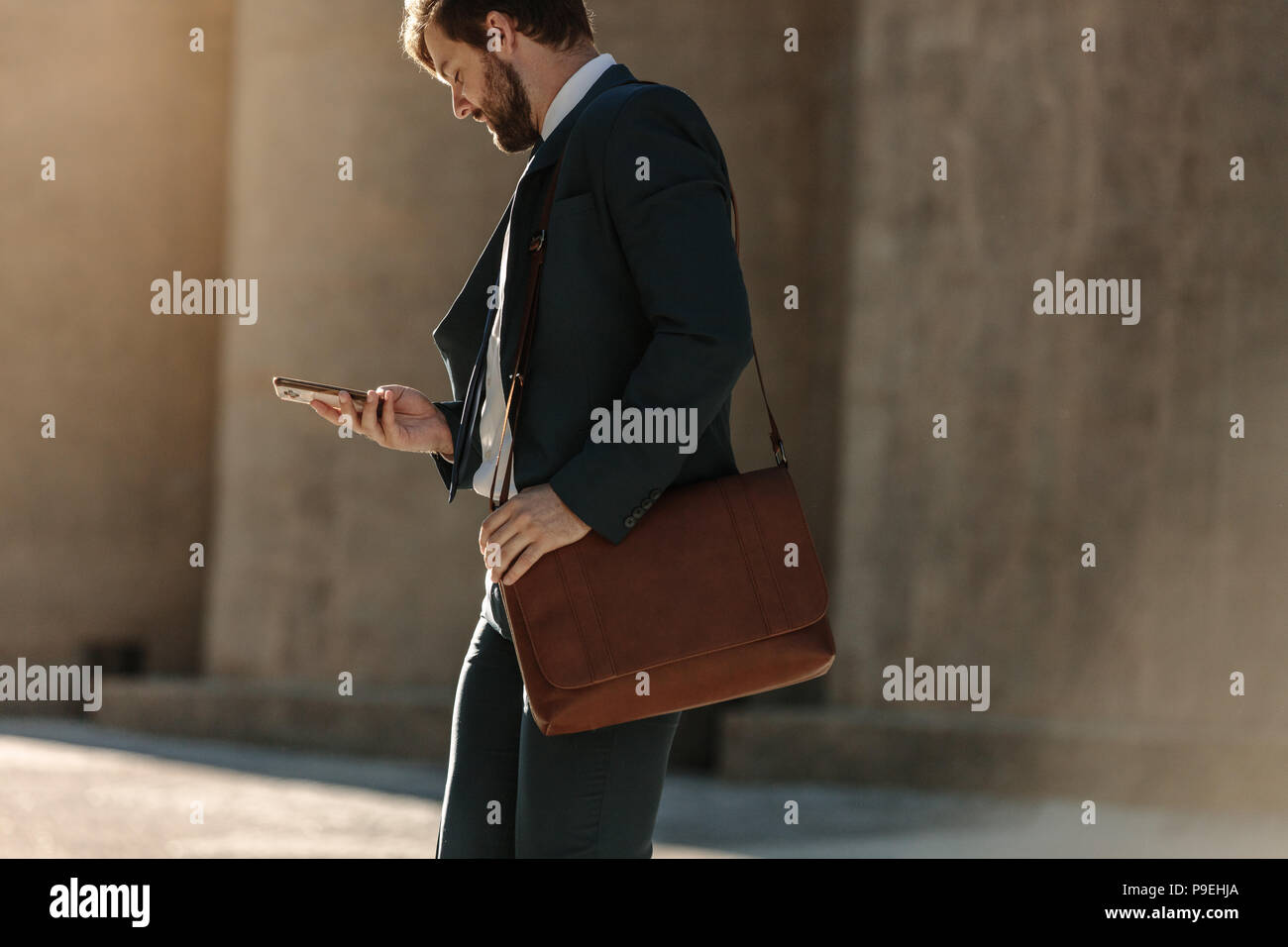 Businessman looking at his mobile phone while walking on street to office. Busy office going man commuting to office carrying his office bag and using Stock Photo