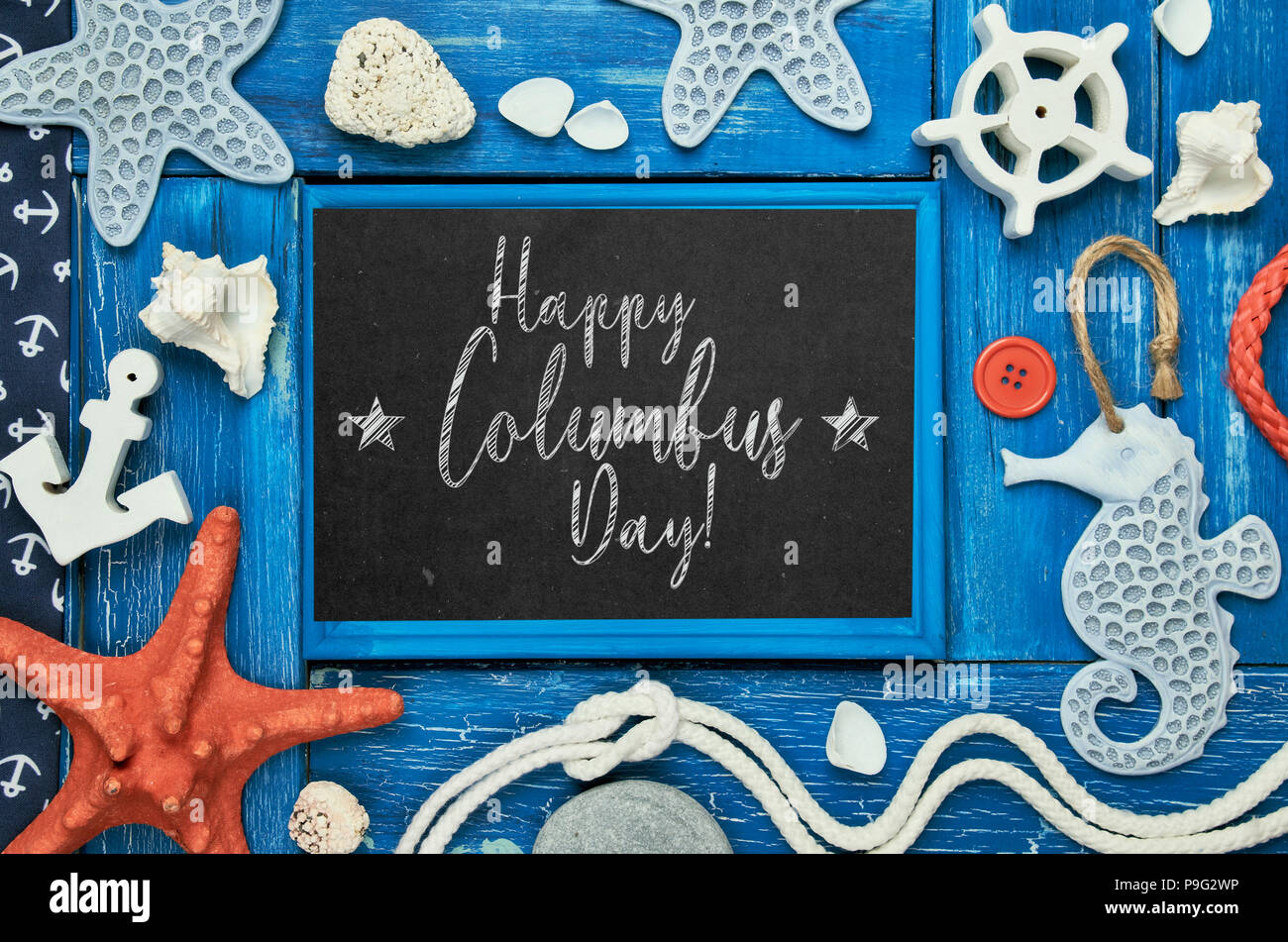 Blank blackboard with sea shells, stones, rope and star fish on blue wooden background, text 'Happy Columbus day' Stock Photo