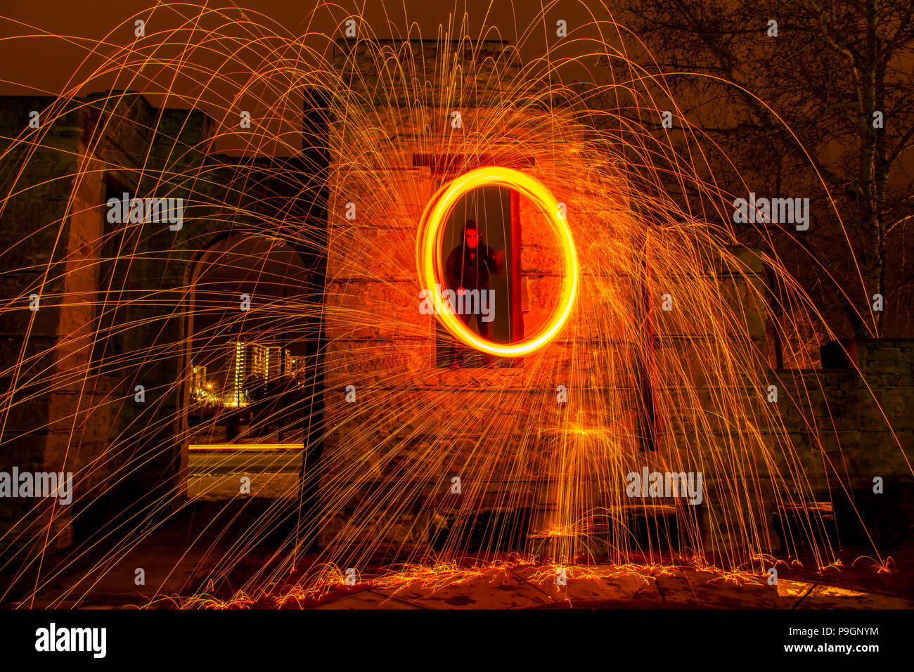 A ring of fire is created from a spinning chunk of burning steel wool on a rope.  A man spins it from a long abandoned stone window. Stock Photo