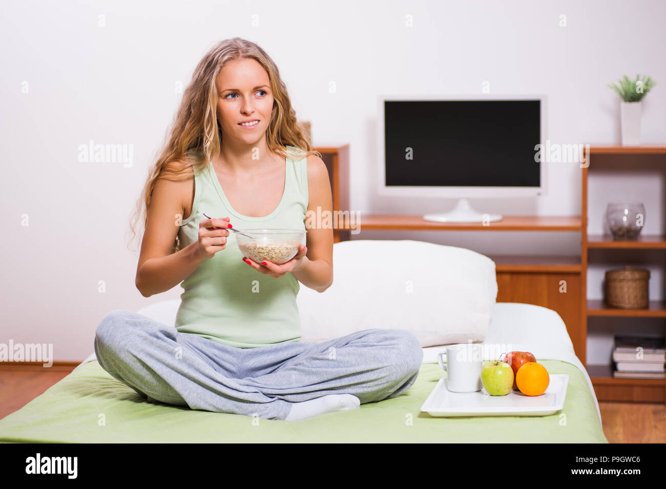 Young woman sitting in her bed and eating oatmeal. It's time for breakfast. Stock Photo