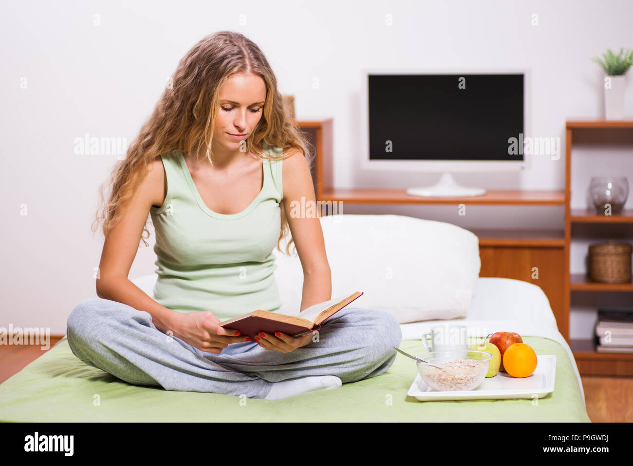 Young woman sitting in her bed and reading book. It's time for breakfast. Stock Photo