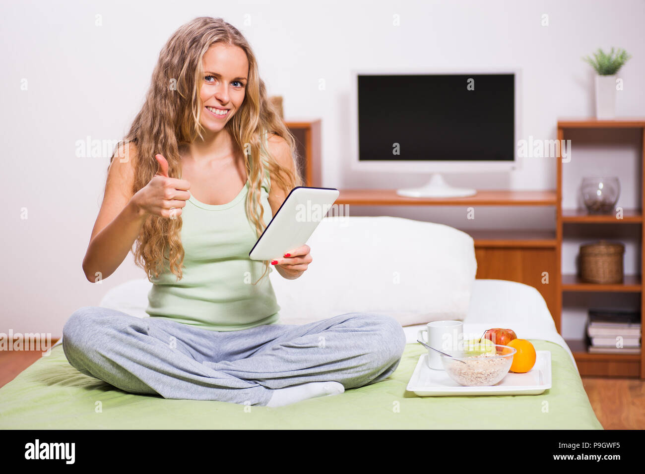 Young woman sitting in her bed and using digital tablet. It's time for breakfast. Stock Photo