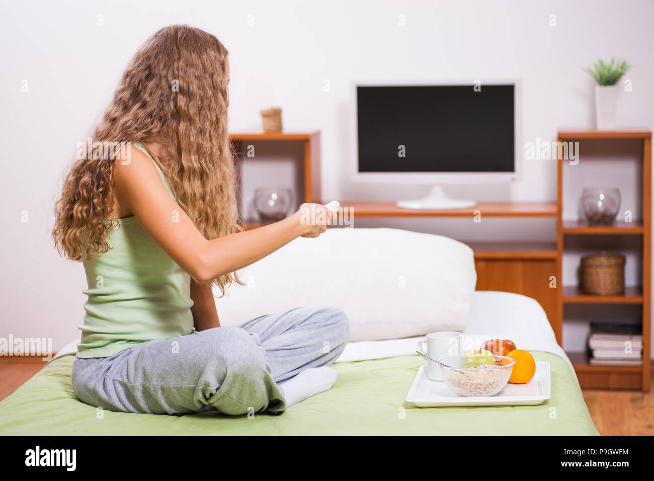 Young woman sitting in her bed and watching TV. It's time for breakfast. Stock Photo