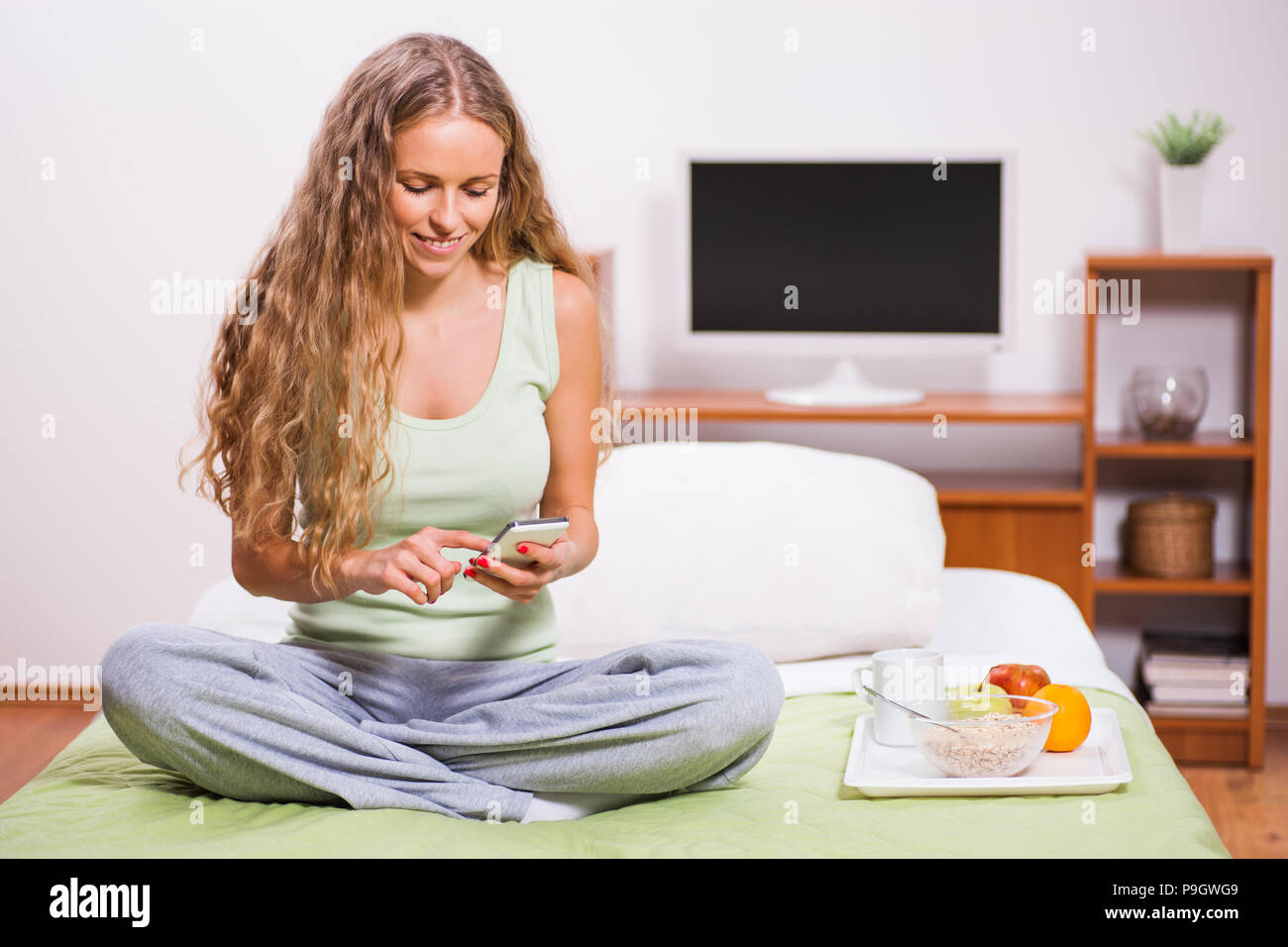 Young woman sitting in her bed and typing on phone. It's time for breakfast. Stock Photo