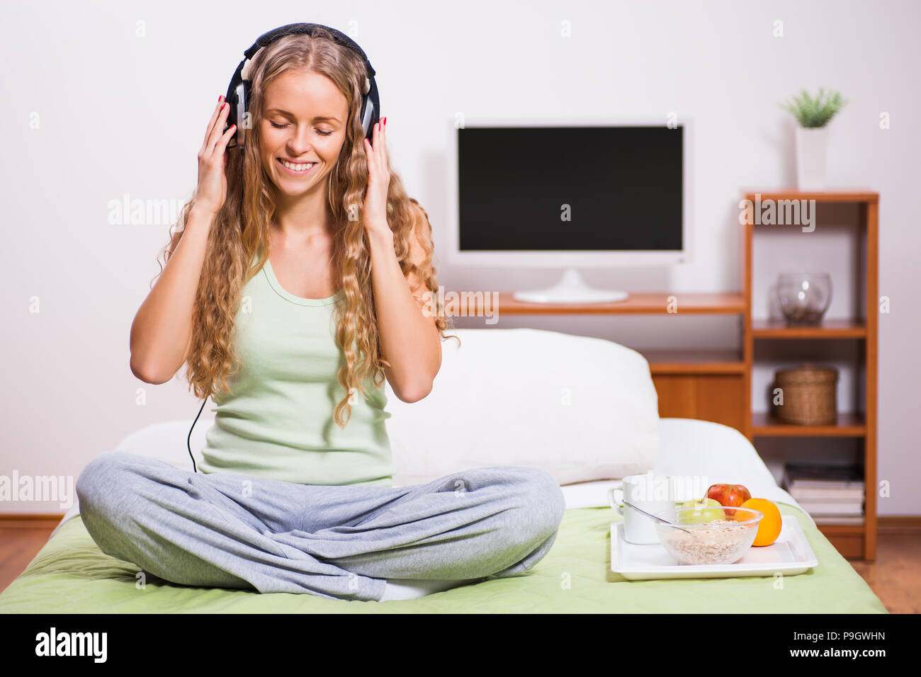 Young woman sitting in her bed and listening music. It's time for snack. Stock Photo