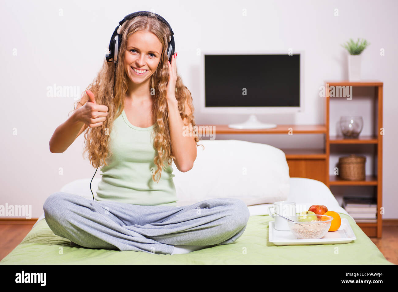 Young woman sitting in her bed and listening music. It's time for snack. Stock Photo